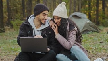 Young happy couple in love tourists sit outdoors in campsite look at laptop screen choose place to travel on electronic map use computer application backpackers communicate laughing enjoy vacation