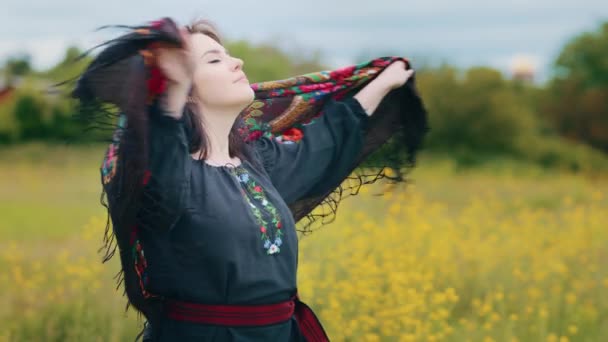 年轻时尚的女人迷人的女孩独自呆在户外穿着传统的乌克兰服装 黑色衣服 手持飘逸的花纹色彩斑斓的围巾站在草地上 披着时尚的围巾 — 图库视频影像