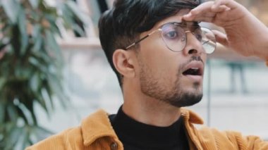 Close-up young indian guy waiting for date meeting puzzled man worries keeps hand on forehead looking out gesture peeking looks into distance with curious expression face waits for friend in search