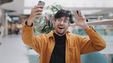Young happy indian guy blogger holds smartphone recording vlog filming video for blog in mall joyful man waving greeting in webcam on phone video conference online takes selfie photo on mobile camera
