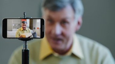 Yaşlı, yaşlı, Kafkasyalı, yaşlı, nüfuzlu iş adamı, çevrimiçi doktor, psikolog, internet öğretmeni tripoda akıllı telefon kullanarak video blogu kaydediyor.