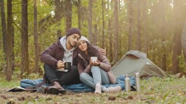 Young hispanic married couple tourists rest in woods camping man and woman in love drinking hot tea talking cute loving guy hugging girl spend nice time together smile communicate travel in nature