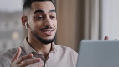 Positive hispanic young man guy male teacher wear wireless earphones look at laptop screen talk at webcam call video chat consult client online provide remote job interview use internet at home office