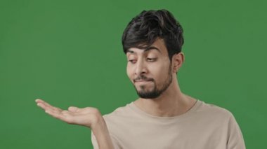 Indian young handsome man stands on green background holds copy space on hand points at empty place arabic smiling brunet guy recommend using mockup blank area for advertisement offer imaginary object