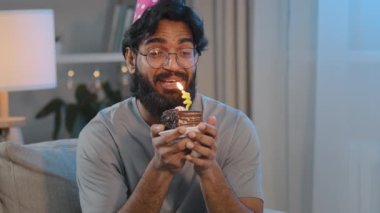 Portrait happy cheerful Arabian Indian bearded man in glasses in festive cap at home evening celebrating anniversary having fun holding small cake making wish talking wishing aloud blowing out candle