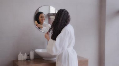 Young happy hispanic woman stands in bathroom in bathrobe looks to mirror combs thick long healthy curly hair using hairbrush feels satisfied morning daily routine advertise haircare treatment concept