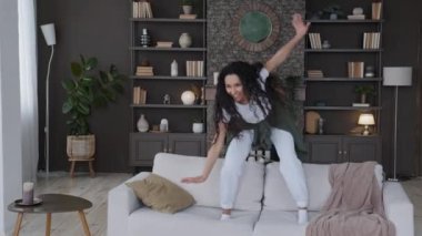 Genç, çekici, sevimli, neşeli bir kadın oturma odasındaki kanepeye atlıyor komik bir şekilde dans ediyor, komik bir şekilde müzik eşliğinde eğleniyor evde tek başına eğleniyor, eğleniyor, komik hareketler yapıyor.