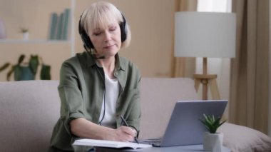 Kulaklıklı kıdemli iş kadını koltukta oturup internetten internet eğitimi alıyor. Webinar uzaktan eğitim alıyor. 60 'lı yılların kadın birinci sınıf kadın sohbet konferansı. Laptop bilgisayarında uzaktan kumandalı yazma notu.