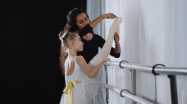 Teen flexible girl kid child ballerina in white tutu stretching leg near barre female adult trainer woman teacher advice learn to dance ballet in dancing school class lesson helps with stretch posture