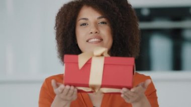Smiling African American girl with curly hair holding giving present showing gift box wrapped in red paper with golden ribbon wishes happy birthday new year Christmas celebration woman congratulations