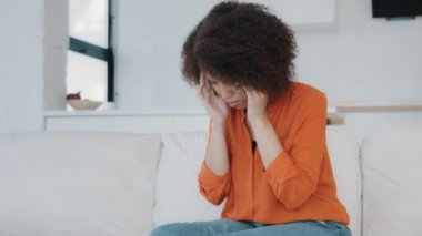 African American girl with curly hair woman sitting at home on couch touching massages temples feels headache dizziness migraine suffers from pain ache holding head stress from illness panic attack