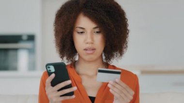 Afro-Amerikalı bir kadın ev mutfağında finansal mobil servis ile online ödeme yapıyor. Kıvırcık saçlı melez kız elinde banka kartı telefonuyla internet ödemeleri yapıyor e-ticaret alışverişi yapıyor.