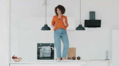 Carefree happy African American woman housewife with curly hair standing on kitchen table dancing alone cooking meal funny cheerful active biracial girl crazy dance energy movement at home having fun