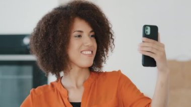 African American girl blogger influencer woman records video blog shooting vlog online streaming chat conference call on web camera mobile phone at home kitchen waving hello talking to smartphone