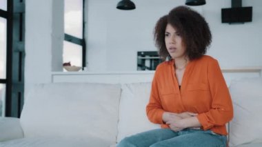 Stresli Afrikalı Amerikalı kadın koltukta oturmuş karnına dokunuyor. Hasta, çift ırklı kıvırcık kız karın ağrısı çekiyor. Mutsuz kadın adet sancısı çekiyor. Acı verici. Karın ağrısından rahatsız.