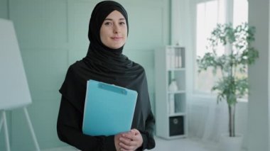 Müslüman Müslüman kadın stajyer kadın modern ofis çalışma alanında elinde belgelerle duruyor. Arap kadın ofis müdürü patron işçi lideri tesettürlü iş kadını kameraya bakıyor