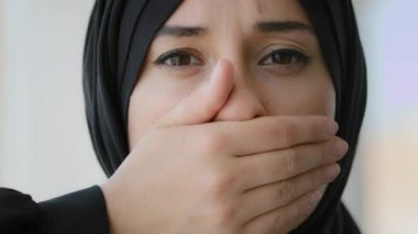 Female portrait indoors Islamic Arabian girl sad afraid Muslim woman Arab lady in black hijab looking at camera covers mouth with hands quiet silence discrimination stop talking oppression of women