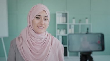 Smiling Arabian Muslim Islamic ethnic business woman influencer recording video blog podcast on smart phone on tripod at home office. Female blogger online teacher shooting mobile vlog webinar calling