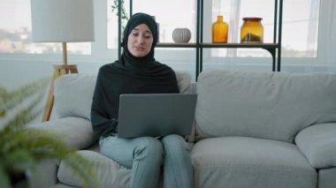 Indian Muslim businesswoman wear black hijab speak look at laptop camera Islamic Arabic lady talk to webcam at home sitting on couch woman has video conference call job interview webinar online chat