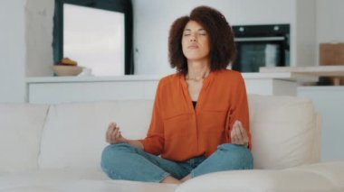 Kıvırcık saçlı, sakin bir Afrikalı kadın nilüfer çiçeği pozisyonunda bacak bacak üstüne atmış bir şekilde oturur. Gözleri kapalı meditasyon yapar, nefes alıp verişi dengeler. Yogi uyumu. Stres yok.