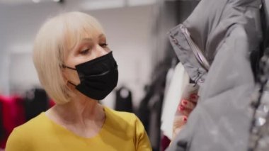 Giysi mağazasında yetişkin bir kadın müşteri koruyucu maskeli beyaz kadın giysi seçiyor. Fiyat etiketine dikkatlice bakıyor. Butik mağazasından moda sonbahar palto alışverişi yapıyor.