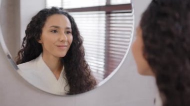 Young calm attractive woman in bathrobe stands in bathroom looks in mirror admiring reflection enjoy healthy skin after uses moisturizing cream facial mask spa procedure natural beauty morning routine