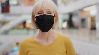 Close-up mature caucasian woman in protective mask standing indoors looking at camera worried serious aware elegant lady in medical respirator protects health from infectious diseases pandemic concept