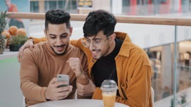 Young happy surprised guy looks at phone screen checks email reads good news win notice won lottery winning bet celebrates victory success excited joyful man supporting friend friendly support concept