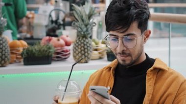 Genç mutlu adam kafede oturur taze sıkılmış meyve suyu içer elinde cep telefonu tutarak sosyal ağdaki uzak arkadaşlarıyla mesajlaşan akıllı telefondan ücretsiz kablosuz ağ kullanır.