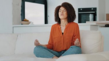 Afro-Amerikalı sakin kadın, oturma odasındaki rahat koltukta nilüfer pozisyonunda oturuyor. Kıvırcık saçlı kız meditasyonu yapıyor. Zihinsel denge meditasyonu yapıyor.