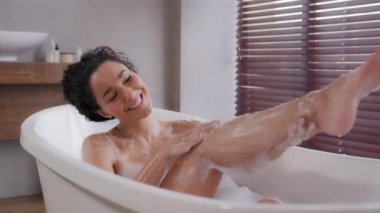 Young happy hispanic woman washing in bath cleans soap body lies in foam bathtub washes feet satisfied girl enjoys daily routine takes care skin relaxing in comfortable bathroom resting alone at home