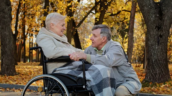 Caucasian senior man holding female hand caring woman with disability on wheelchair husband accompanies sick wife during rehabilitation in autumn park grandparents couple spend time together outdoors