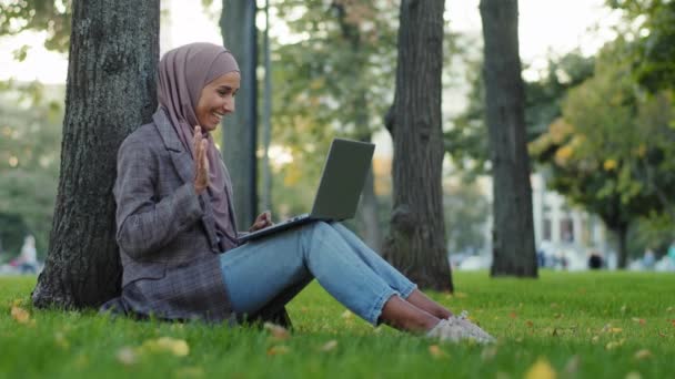 ハッピーイスラム教徒のイスラム教徒の女の子ビジネス女性でヒジャーブ市内公園で緑の芝生の上に木の近くに座っている友人とチャット家族のリモート会話オンラインビデオチャット電話会議とノートパソコンの話 — ストック動画