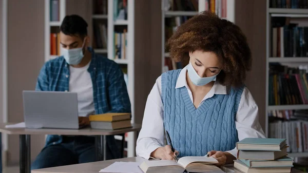 Students study in library in protective mask comply with quarantine measures young clever girl student reading book looking for information in textbook writes notes preparing for exam doing homework