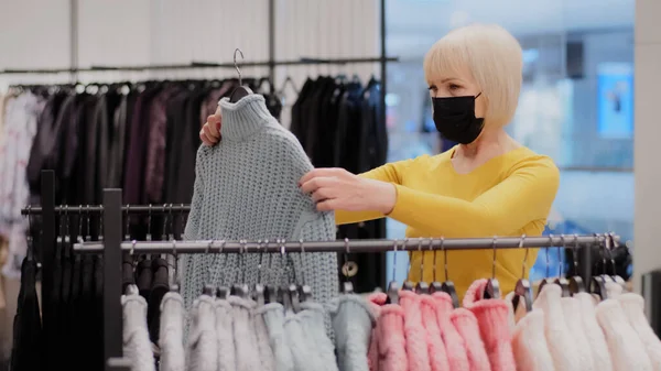 Mature focused woman shopper in protective mask searching clothes in boutique attractive middle-aged lady customer in clothing store looking for new outfit in search quality sweater on sale discount