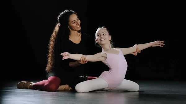 Hispanic woman coach dance trainer teacher with long curly hair help girl teenager bend back holding hands at side sitting on floor ballerina gymnast stretching advice trains position ballet class