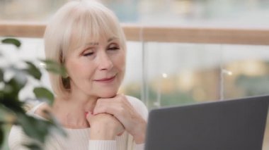 Female portrait close-up cute attractive mature woman holding hands under chin looking at camera smiling happy caucasian elderly businesswoman sitting at workplace thoughtfully looks at laptop screen