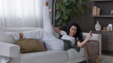 Genç, kaygısız, oyuncu bir kadın, rahat bir kanepede uzanıyor, hafta sonunun tadını çıkarıyor, eğleniyor, eğleniyor, kanepede akrobatik hareketler yapıyor, komik danslar yapıyor, şehvetli hareketler yapıyor, kıvırcık saçlar sallıyor.