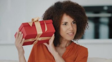 Merak uyandıran mutlu doğum günü kızı kıvırcık saçlı Afrikalı kadın hediye paketini sallıyor ve evde kulaklarında bayram kutlaması olduğunu düşünerek hediyesini dinliyor. Yüksek