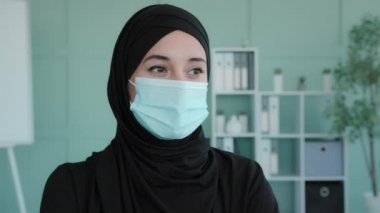 Portrait Arab girl Islamic Muslim woman wearing black hijab wearing medical mask in office businesswoman in covid quarantine coronavirus epidemic indoors looking away thinking dreaming contemplating