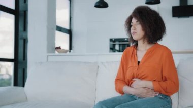 Üzgün Afrikalı kadın, evde kıvırcık saçlı, hasta kız, mutfakta koltukta oturuyor, karnına dokunuyor, karnına dokunuyor, karın ağrısı çekiyor, acı çekiyor, midesi ağrıyor, hazımsızlık çekiyor.