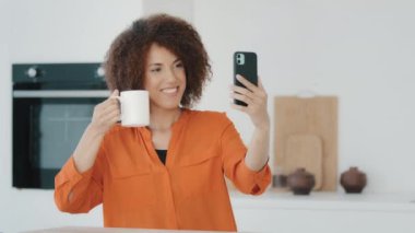 African American girl blogger woman influencer holding cup of tea coffee shows to camera makes photo records video blog online call with mobile phone win victory reaction web winning at home kitchen
