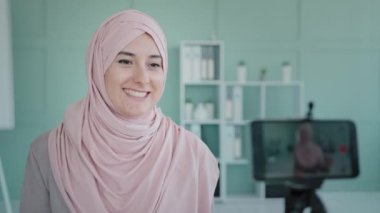 Distant teacher mentor student Arabian Muslim Islamic businesswoman influencer girl in hijab recording video blog podcast online lesson class phone on tripod waving hello mobile remote vlog telesales