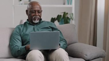 African American bald bearded businessman teacher writer entrepreneur checking data online use computer application connection wifi network at home office sitting on comfortable sofa typing on laptop