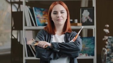Kafkas kızı kendine güvenen sanatçı kadın ressam boya fırçası spatulasını boyamak için elinde ekipman olan resim çizdikten sonra resim stüdyosunda poz veren kırmızı saçlı kadın ressam.