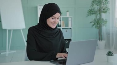 Gülümseyen mutlu İslam iş kadını, siyah tesettürlü, İslami Arap kız öğrenci, Müslüman kadın, dizüstü bilgisayarla çalışıyor, internette sohbet ediyor, bilgisayar e-ticaret uygulaması kullanıyor.