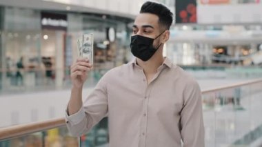 Arap İspanyol işadamı şık giyinmiş tıbbi maskeli adam alışveriş merkezinde karantinada ve indirimli kameraya bakan para banknotlarıyla.
