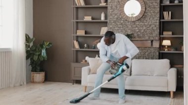 Heyecanlı, olgun, olgun bir adam mutlu dans eden, yetişkin, Afrikalı, bekar ev sahibi, elektrikli süpürgeyle oturma odasında ev işleri yapan, aktif ev işleri yapan, ev işleri yapan,