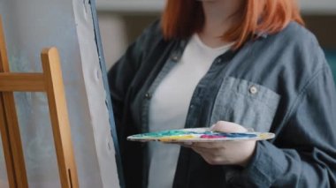 Close-up red-haired girl artist inspired female painter woman with palette and brush paints on canvas with easel create picture with acrylic colors oil paint drawing small business painting talent art