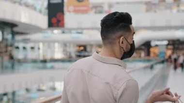 Maskeli, Hint asıllı, siyah tıbbi maskeli İspanyol bir adam alışveriş merkezinde erkek müşteri alıcısı elinde alışveriş çantası hediye paketleri, elinde kamera indirimleri var.